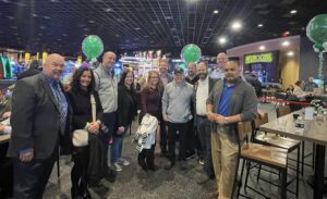A group of United Regional Chamber of Commerce members posing together at the Wahlburgers ribbon-cutting event inside Plainridge Casino in Plainville, MA. The indoor venue is lively with neon lights, green balloon arrangements, and casino arcade decor in the background.